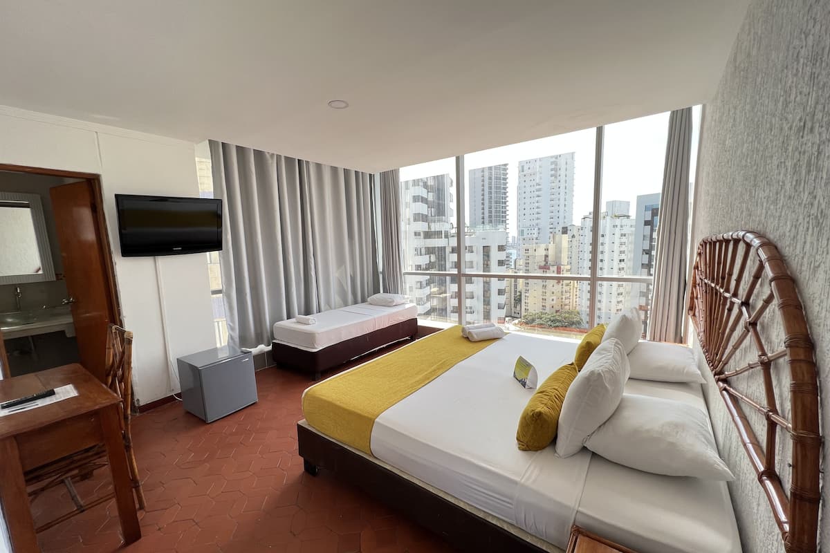A hotel room with a bed, a desk, a chair, a TV, and a large window with a city view.