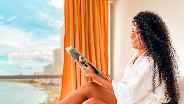 Una persona sentada en una cama en una habitación con vista a un cuerpo de agua y un paisaje urbano.