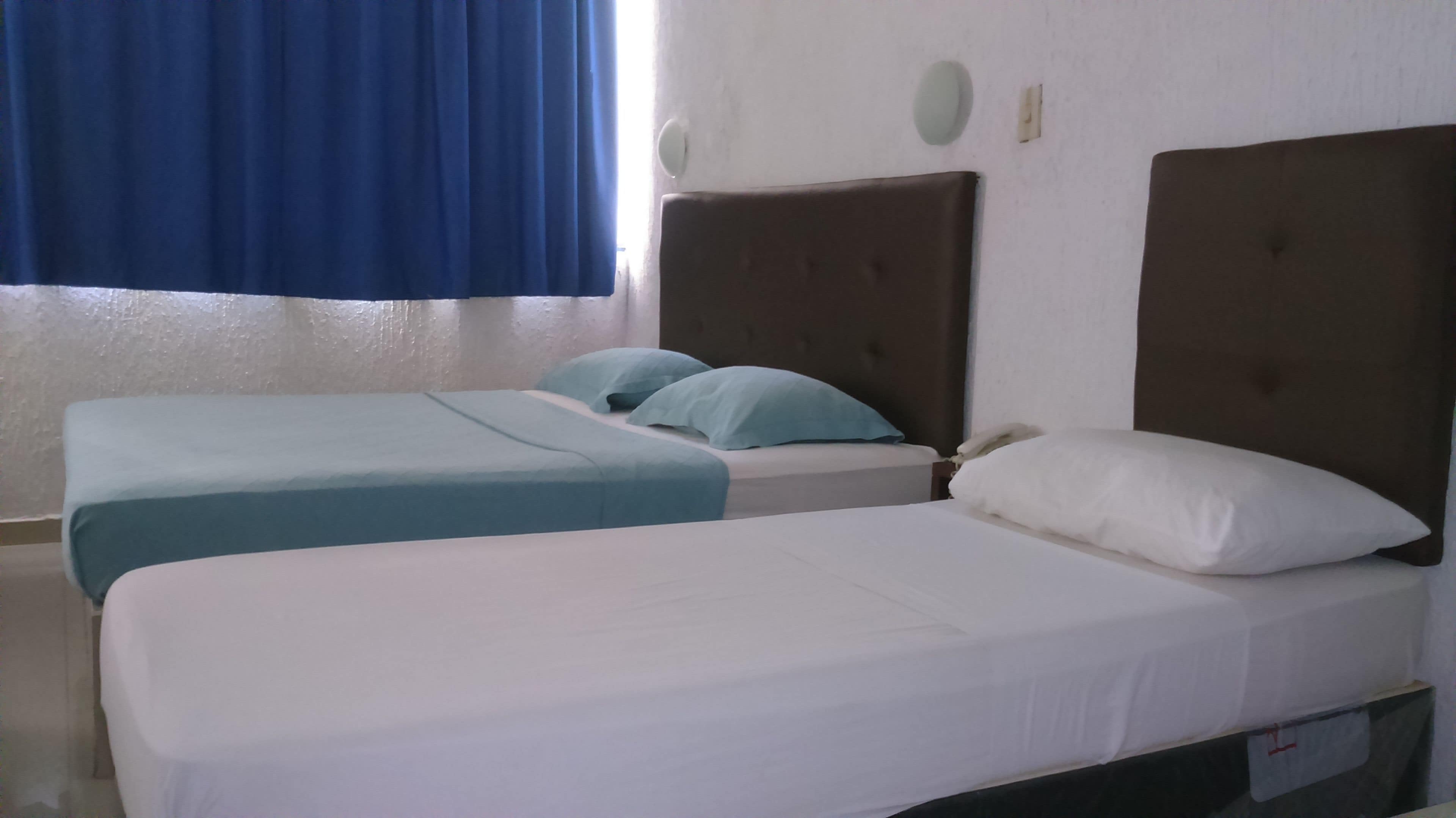 A hotel room with two beds, one with a white mattress and the other with a blue mattress. There is a brown headboard on the right side of the room.
