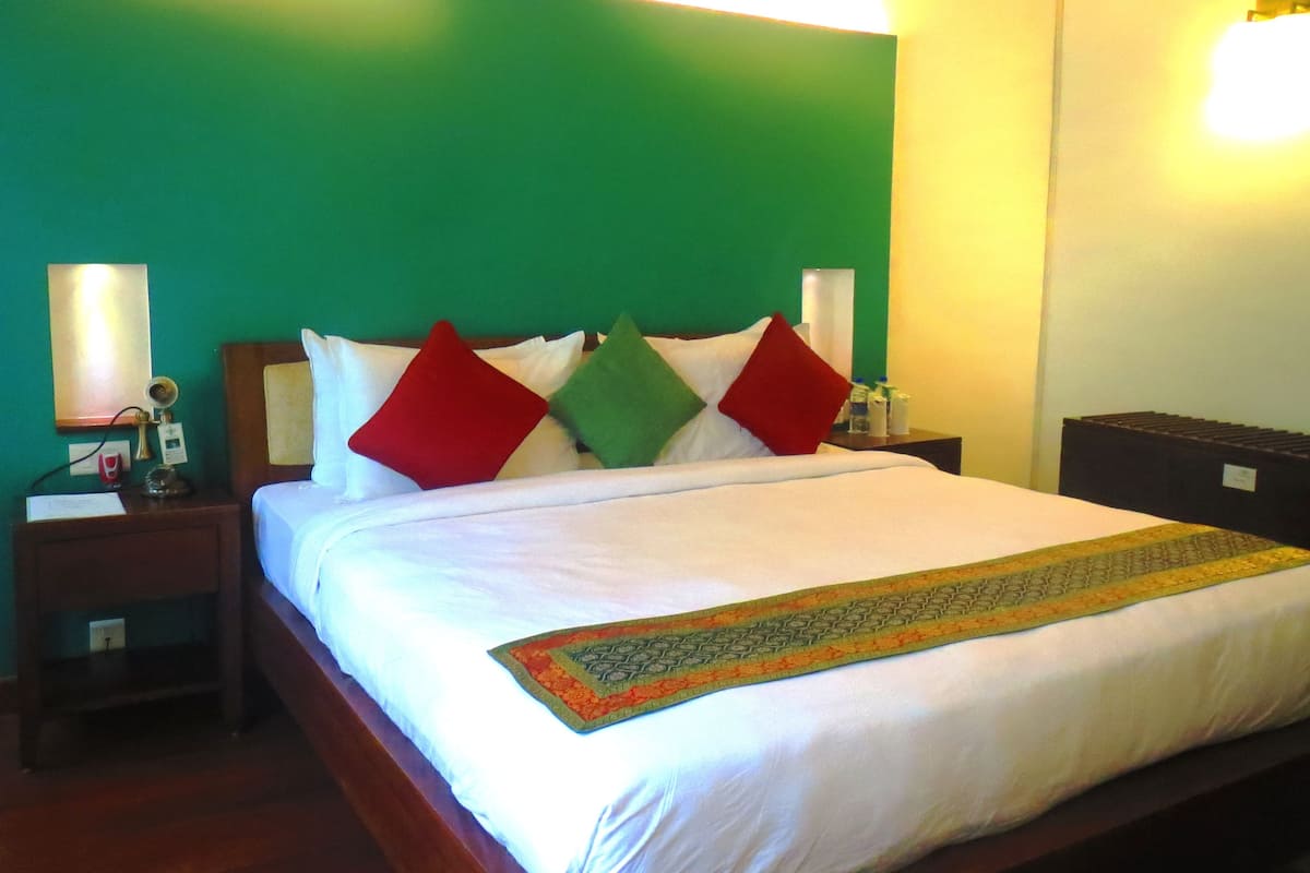 Una cama bien hecha con cubrecama blanco y una colcha decorativa, una mesita de noche con lámpara y teléfono, y una pared verde con una luminaria.