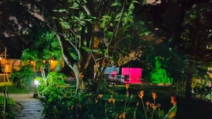 Un jardín nocturno con un sendero de piedra, abundante vegetación y una estructura iluminada.