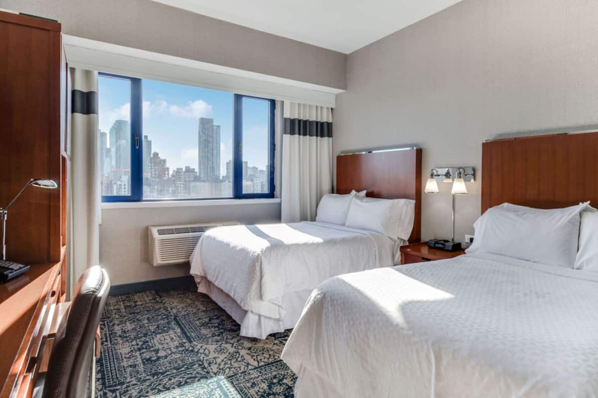 A hotel room with two beds, a large window with a city view, a desk with a lamp, and a patterned rug.