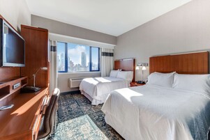 Ein Hotelzimmer mit zwei Betten, einem Schreibtisch, einem Fernseher und einem großen Fenster mit Blick auf die Stadt.