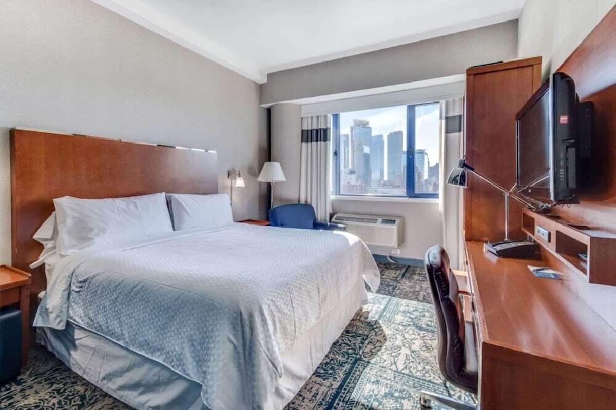 A hotel room with a large bed, a desk with a computer, a chair, a TV, and a window with a city view.
