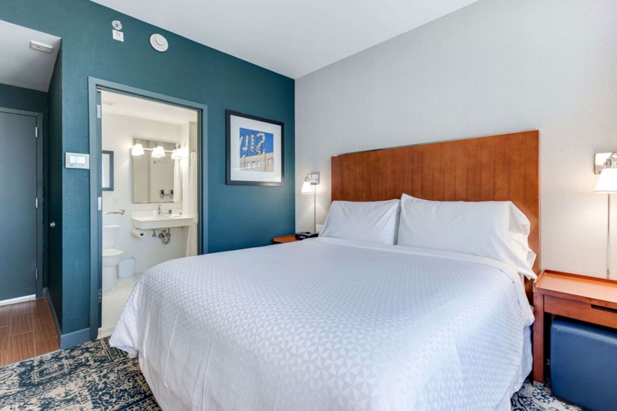 A hotel room with a large bed, a wooden headboard, a nightstand, a bedside lamp, a bathroom with a mirror, and a framed picture on the wall.