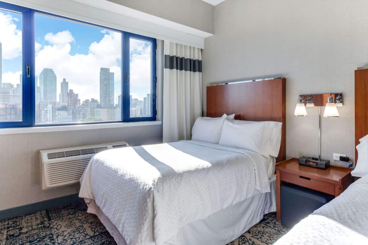 A hotel room with a bed, a nightstand with a lamp, a window with a city view, and a wall-mounted heater.