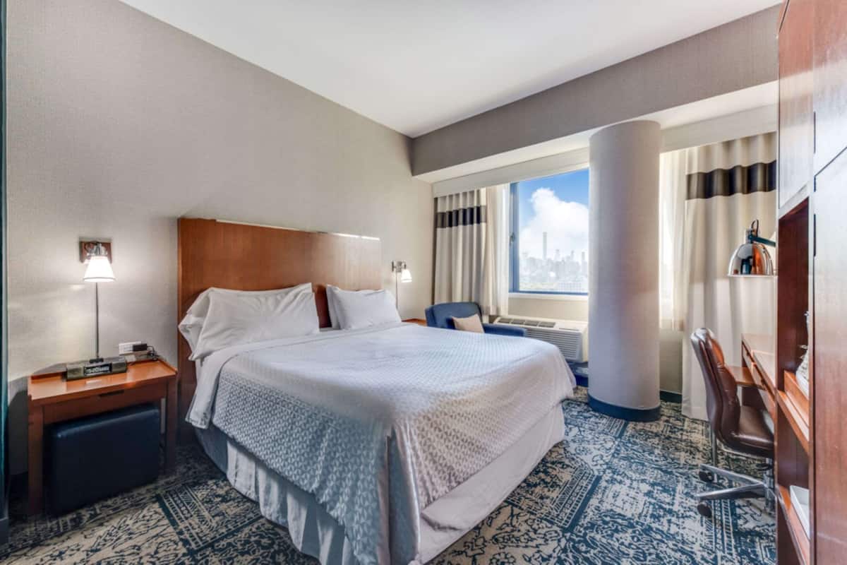 A hotel room with a large bed, a desk, a chair, a nightstand, a lamp, a window with curtains, and a city view.