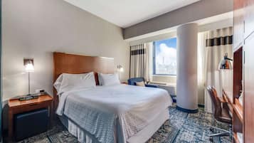 A hotel room with a large bed, a desk, a chair, a nightstand, a lamp, a window with curtains, and a city view.