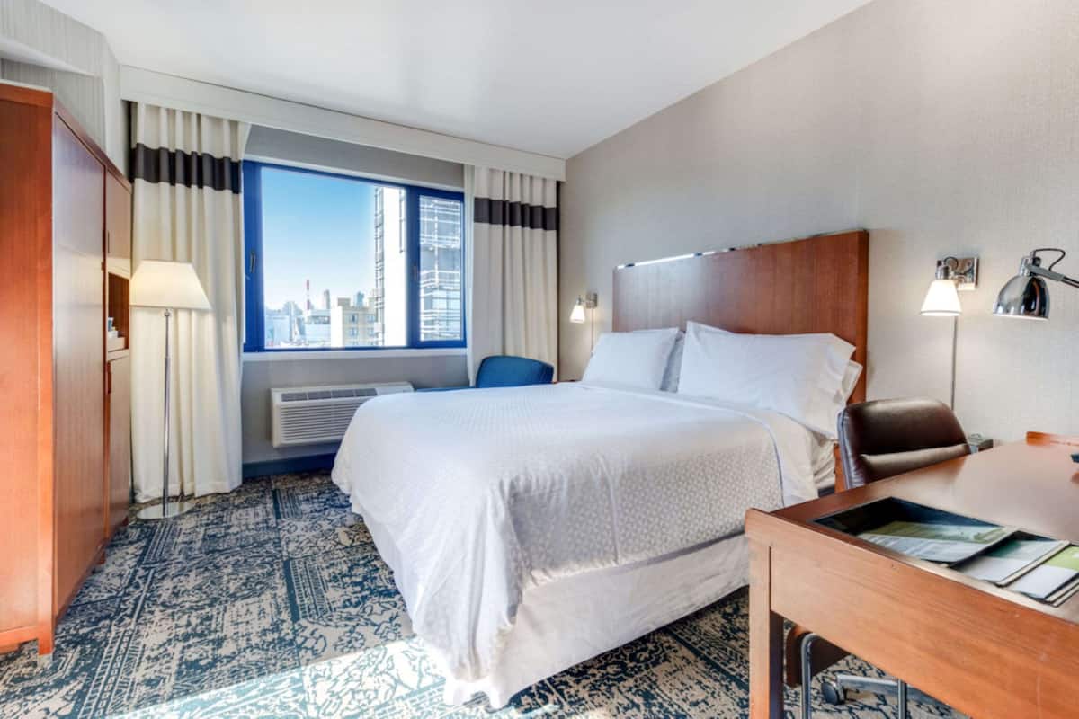 A hotel room with a large bed, a desk, a chair, a window with a city view, and a patterned carpet.