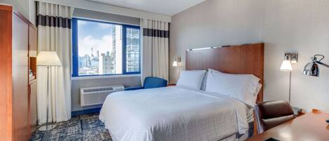 A hotel room with a large bed, a desk, a chair, a window with a city view, and a patterned carpet.