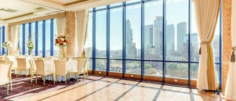 A spacious room with a large window offering a city view, a long table set for dining, and elegant decor.