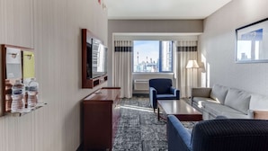 Habitación de hotel con vista a la ciudad, televisor de pantalla plana, sofá, sillón, mesa de centro y escritorio con botellas de agua.