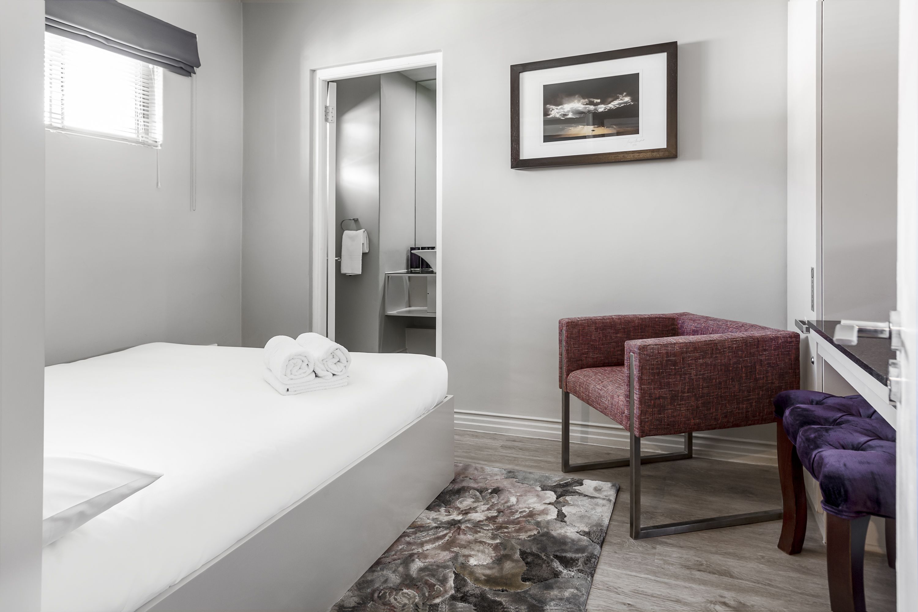 Una camera d'albergo con un letto, una sedia, un tavolino, uno specchio e un quadro appeso al muro.