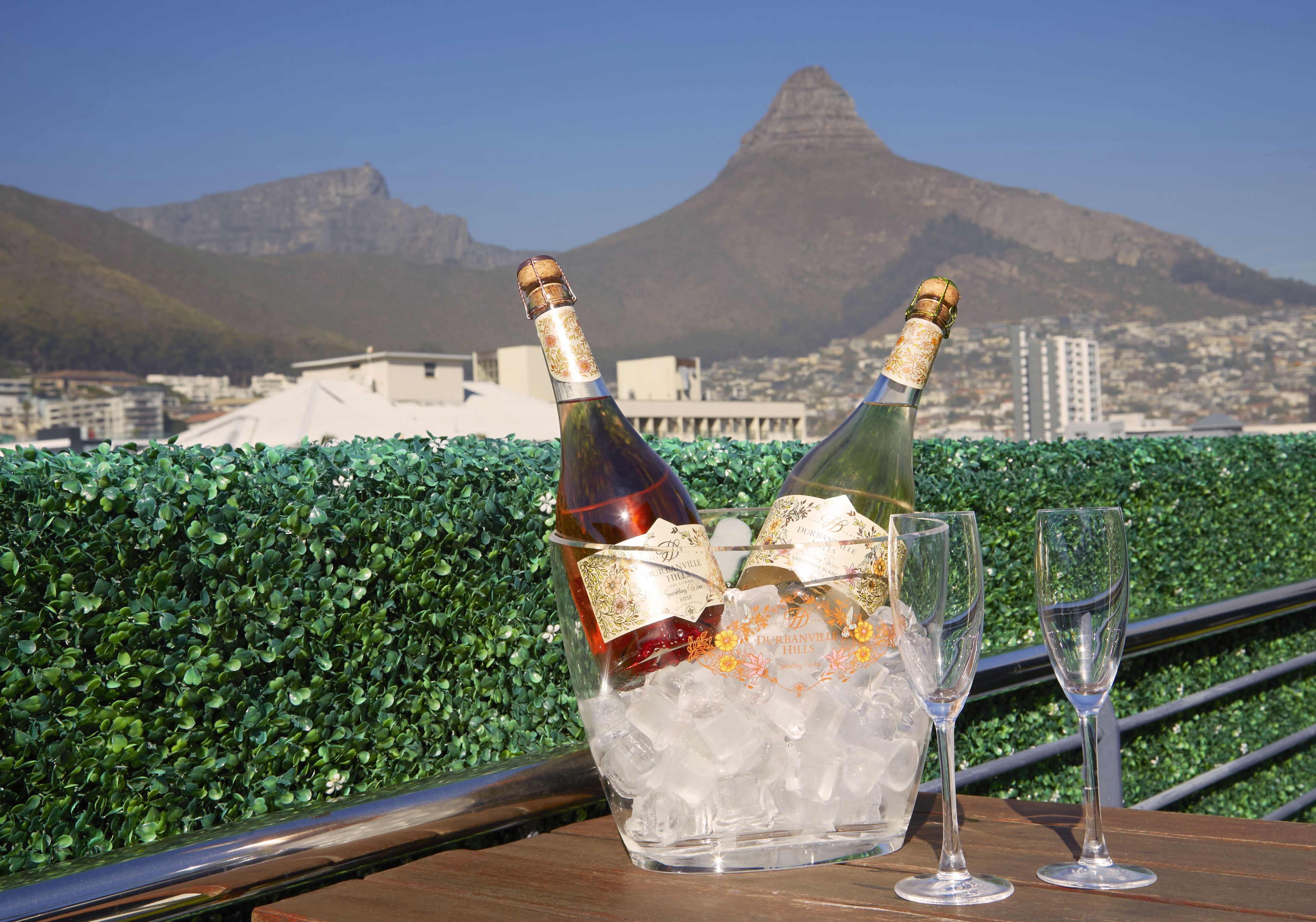 Een tafel met twee champagneflessen en twee glazen, geplaatst voor een achtergrond van een stadsgezicht en bergen.