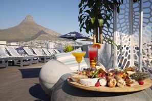 A dining table with a variety of foods and drinks, a beach view, and lounge chairs in the background. - The Hyde All-Suite Hotel (Cape Town)
