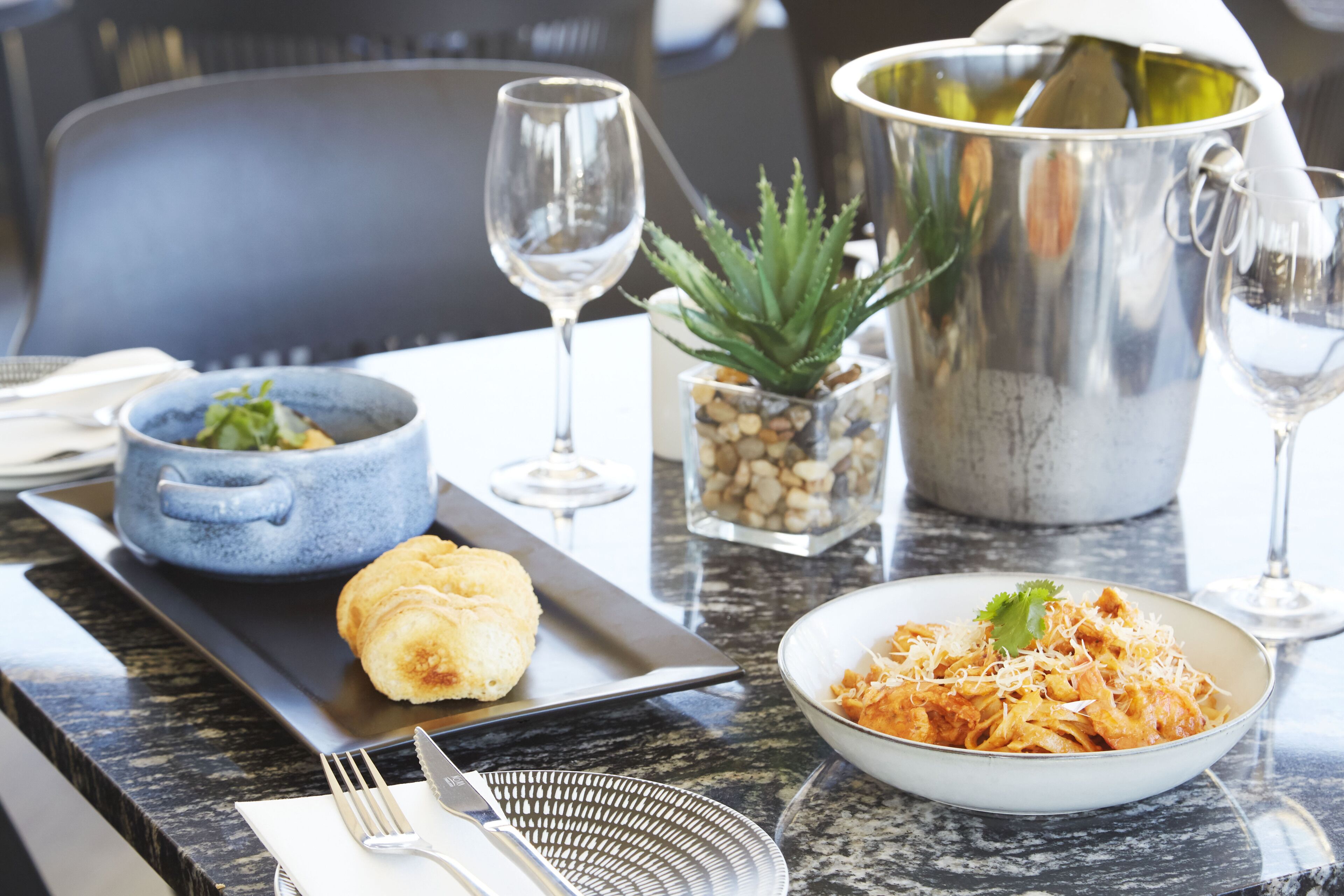 Une table à manger avec une assiette de pâtes, un petit bol de légumes verts, une pâtisserie et un verre à vin.