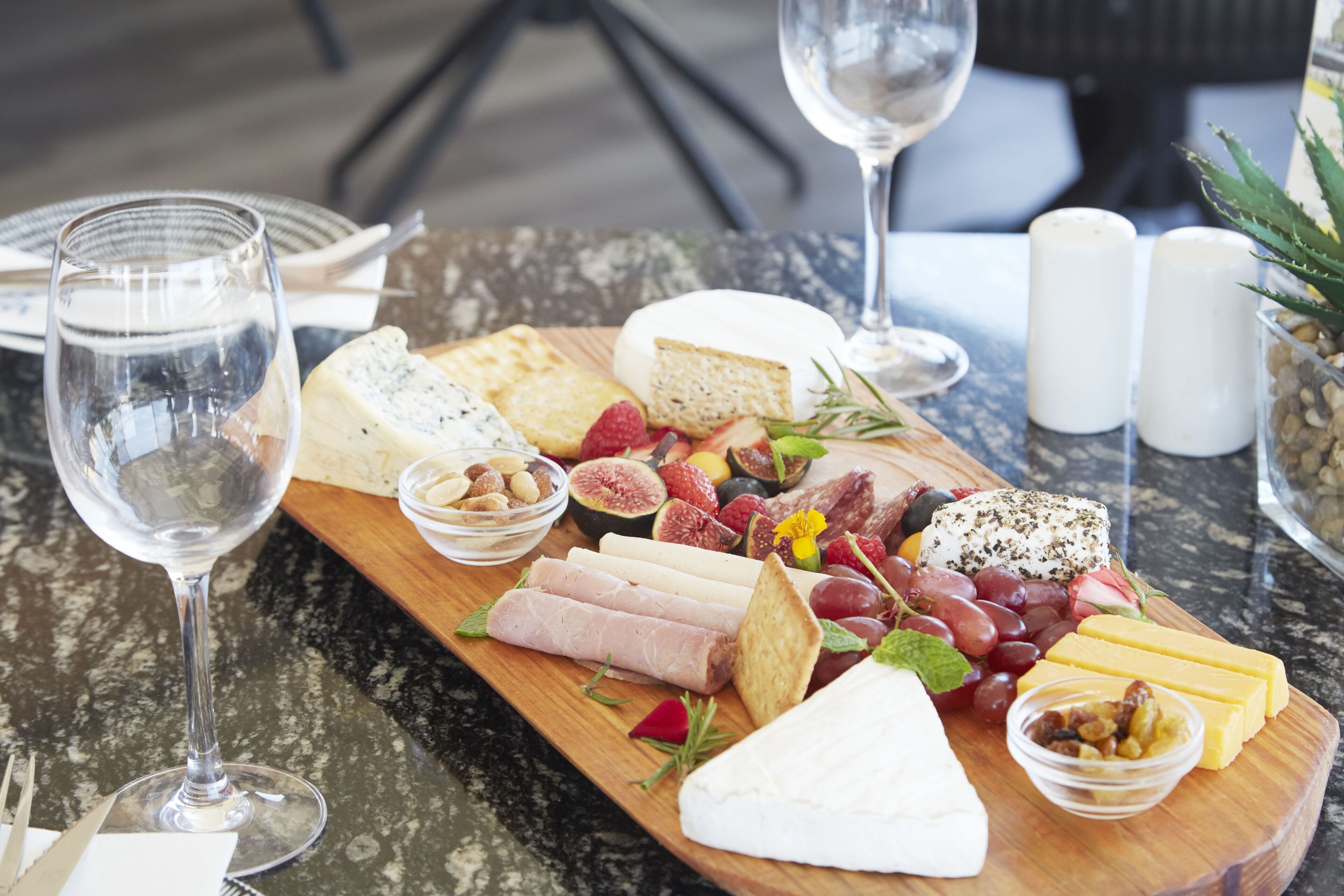 A charcuterie board with various cheeses, fruits, and nuts on a wooden board, accompanied by wine glasses and salt and pepper shakers.