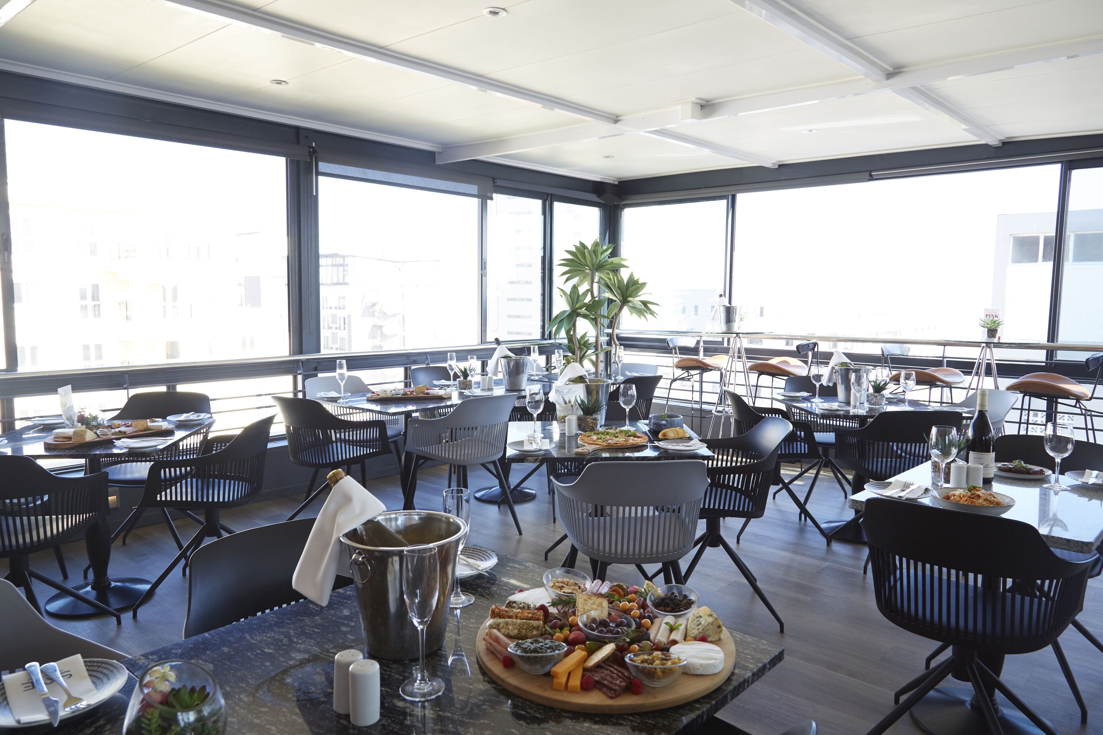 A spacious dining area with multiple tables and chairs, large windows, and a variety of food and drink items on the tables.