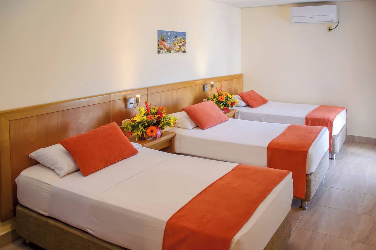 Une chambre d’hôtel avec trois lits, chacun doté de draps blancs et de touches de couleur orange. On y trouve une tête de lit en bois, un climatiseur fixé au mur et un petit tableau au-dessus des lits.