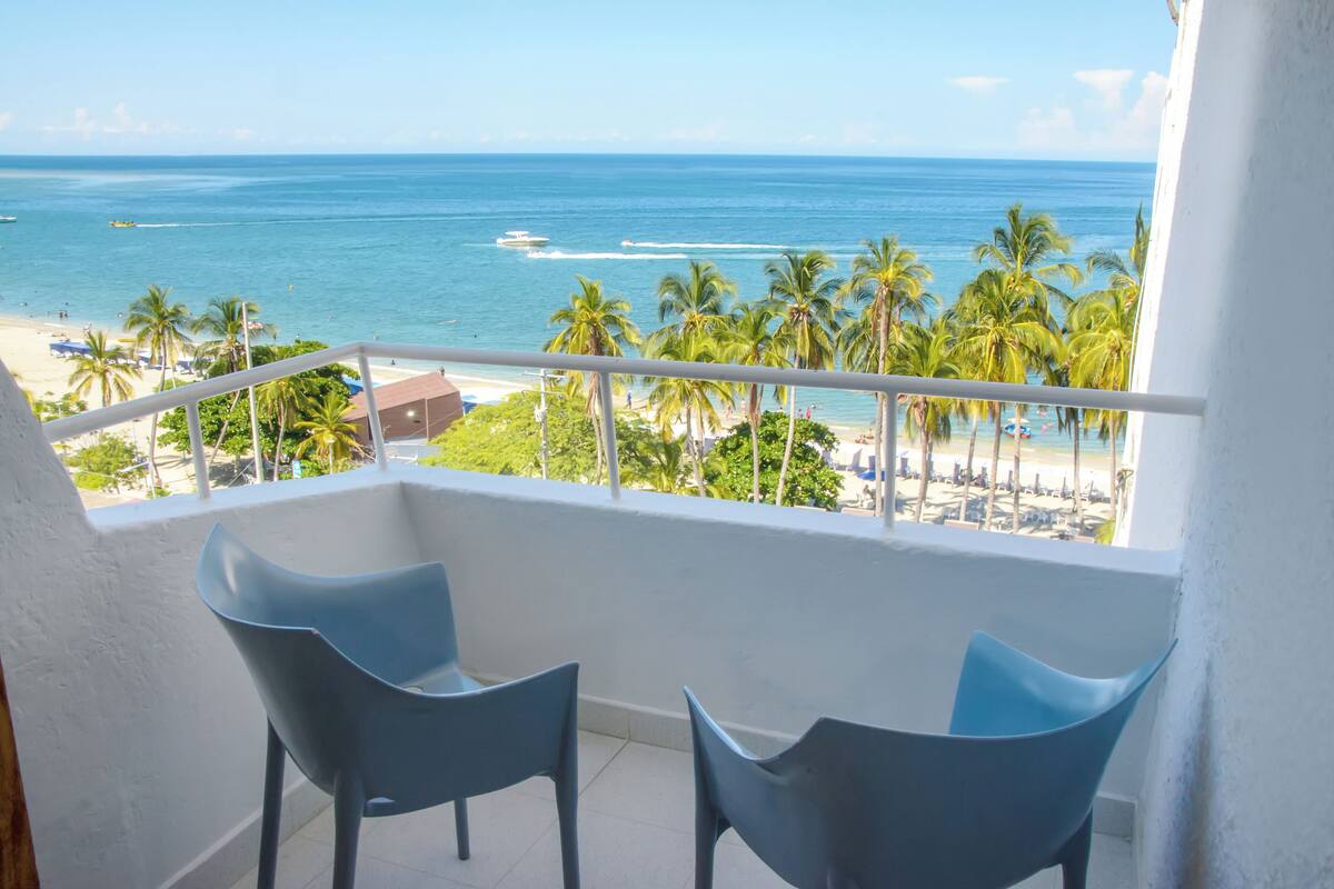 Un balcon avec deux chaises bleues, donnant sur une plage bordée de palmiers et sur l’océan.