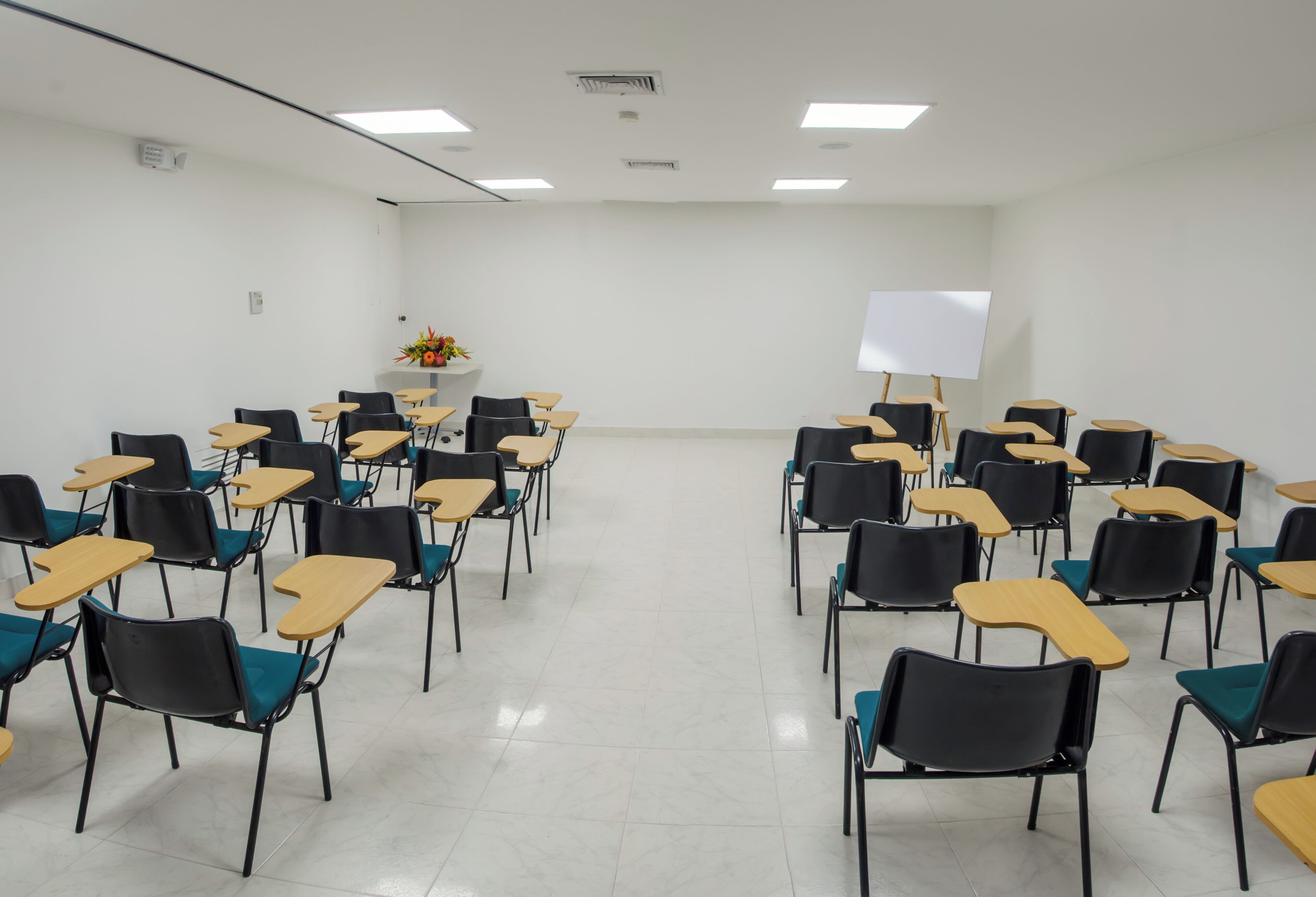 A classroom with rows of desks and chairs, a whiteboard, and a flower arrangement on a shelf.