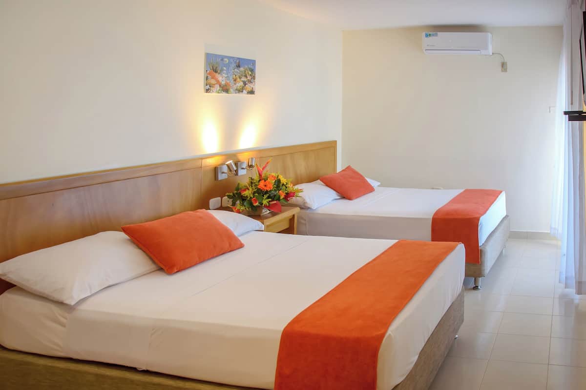 Deux lits simples avec des couvre-lits orange, une table de chevet ornée d’un vase de fleurs et un appareil de climatisation fixé au mur.