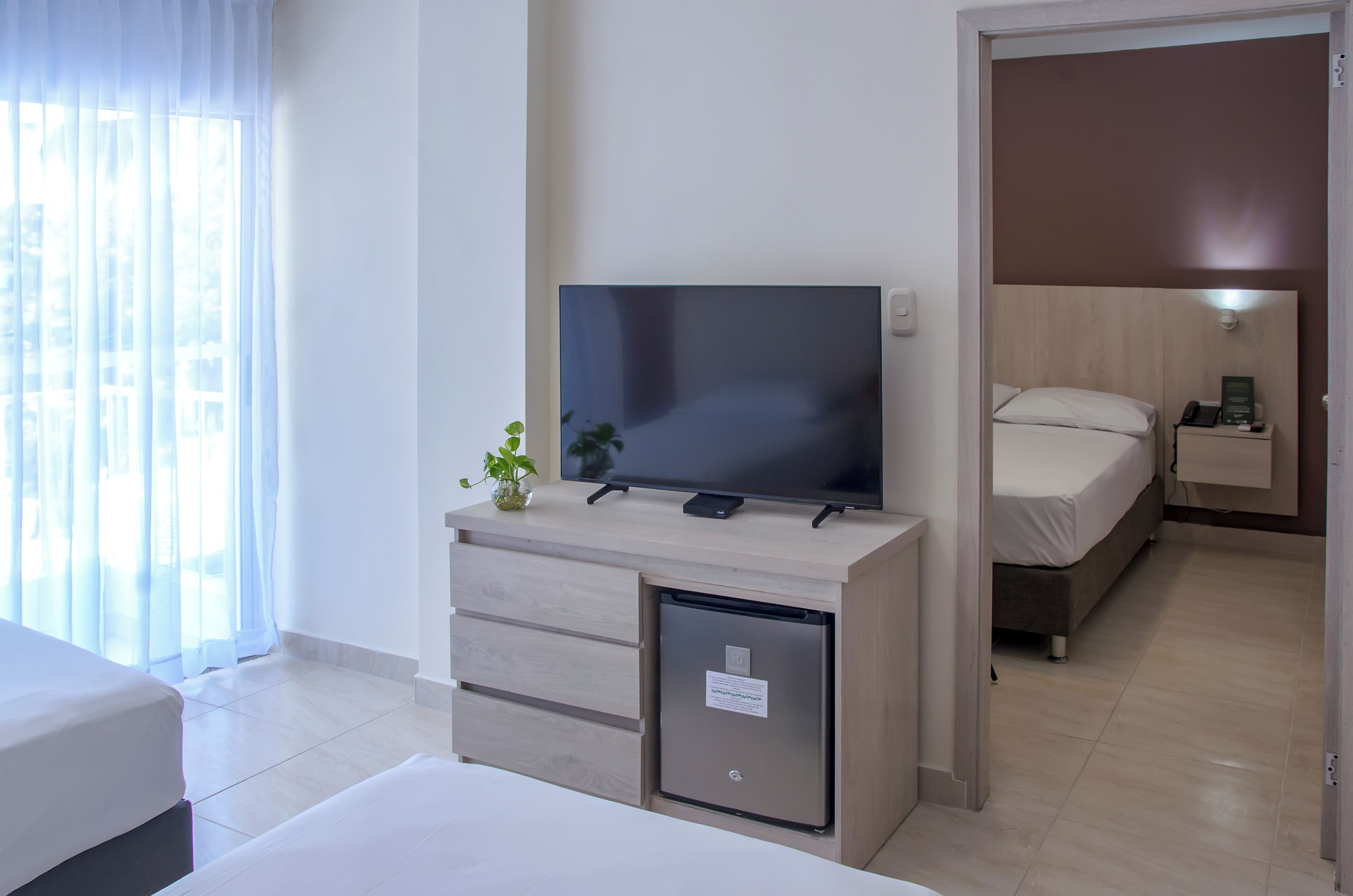 Une chambre d’hôtel avec un lit, un téléviseur posé sur un bureau en bois, un petit réfrigérateur et une table de chevet avec une lampe.