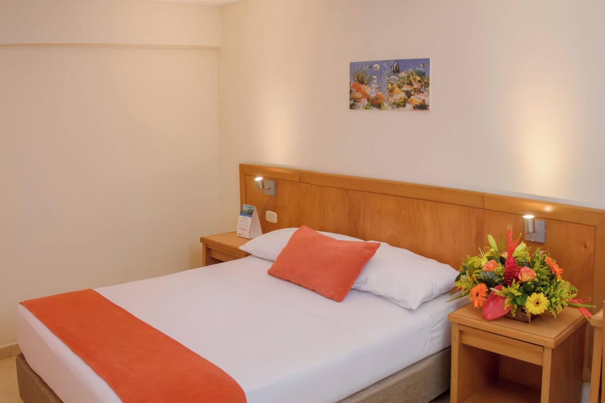 Une chambre d’hôtel comprenant un lit, une table de chevet, une petite table de nuit agrémentée d’un arrangement floral et une œuvre d’art murale.