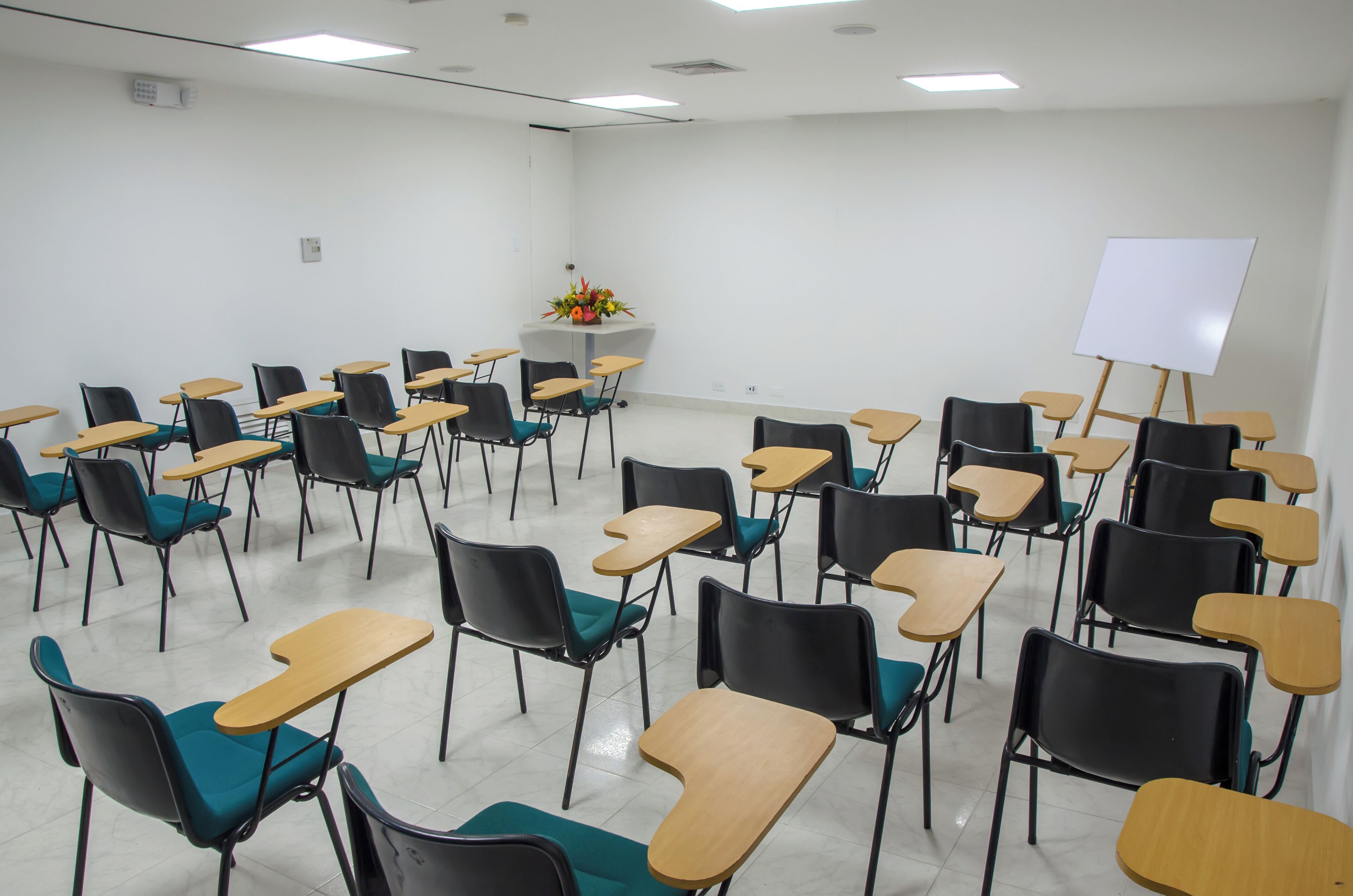 A classroom with rows of desks and chairs, a whiteboard, and a flower arrangement on a podium.