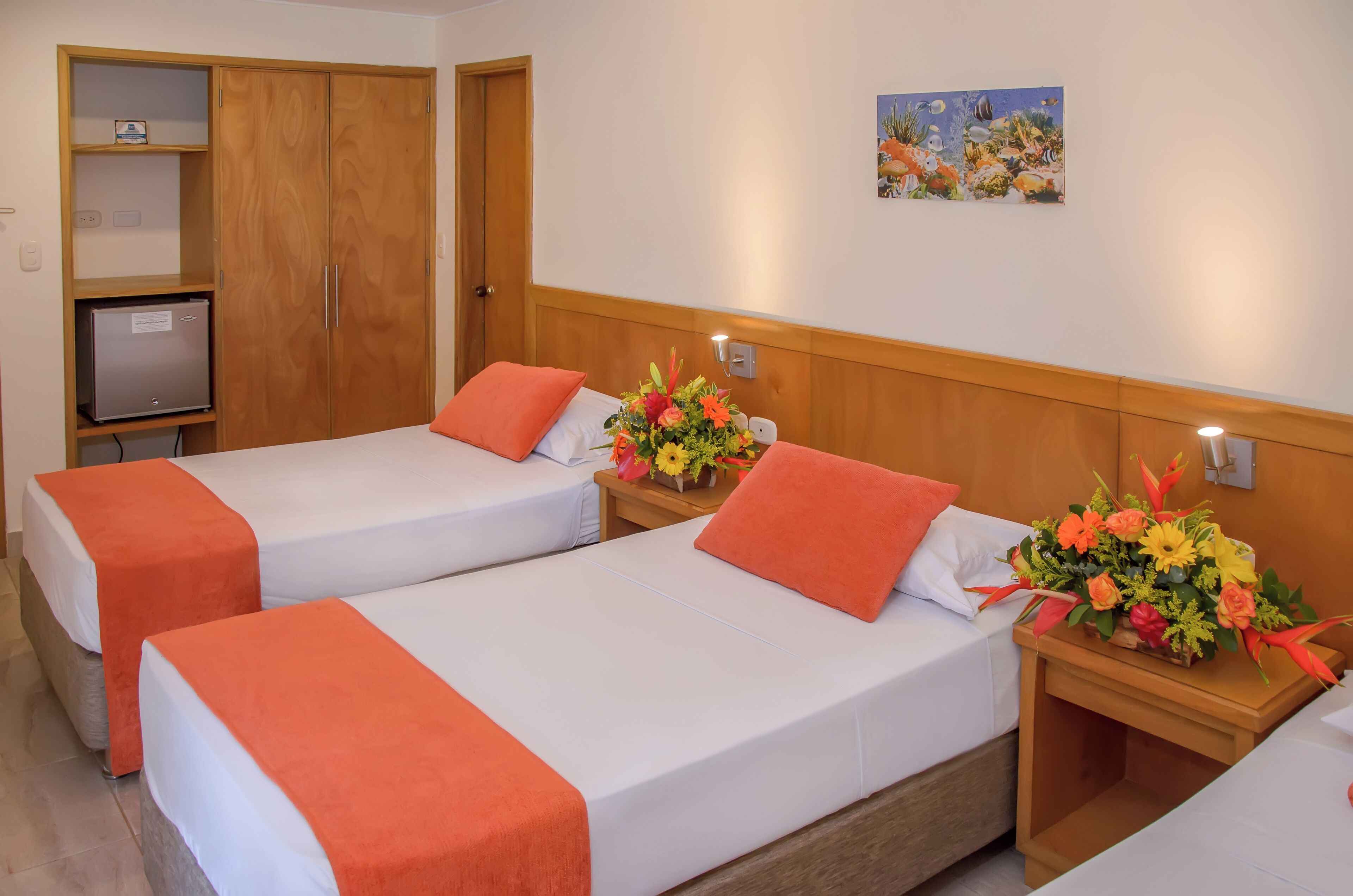 A hotel room with two beds, each with an orange bedspread and pillow, a wooden wardrobe, a small desk with a lamp, and a wall-mounted painting.