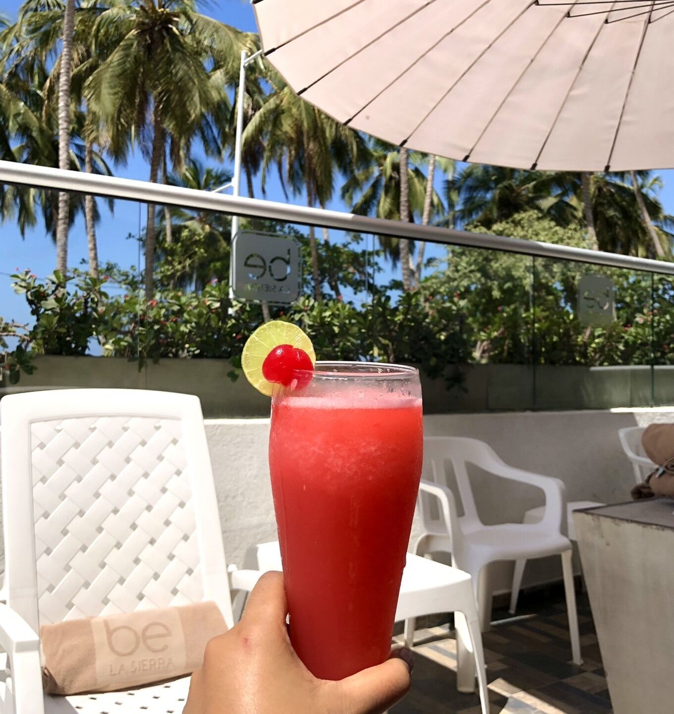 A hand holding a red drink with a cherry garnish, a white chair, and a white umbrella in a sunny outdoor setting.