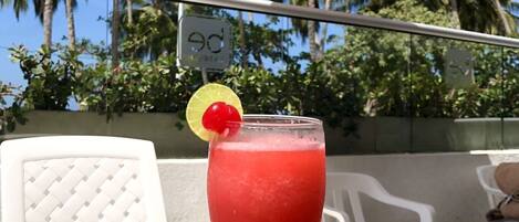 A hand holding a red drink with a cherry garnish, a white chair, and a white umbrella in a sunny outdoor setting.
