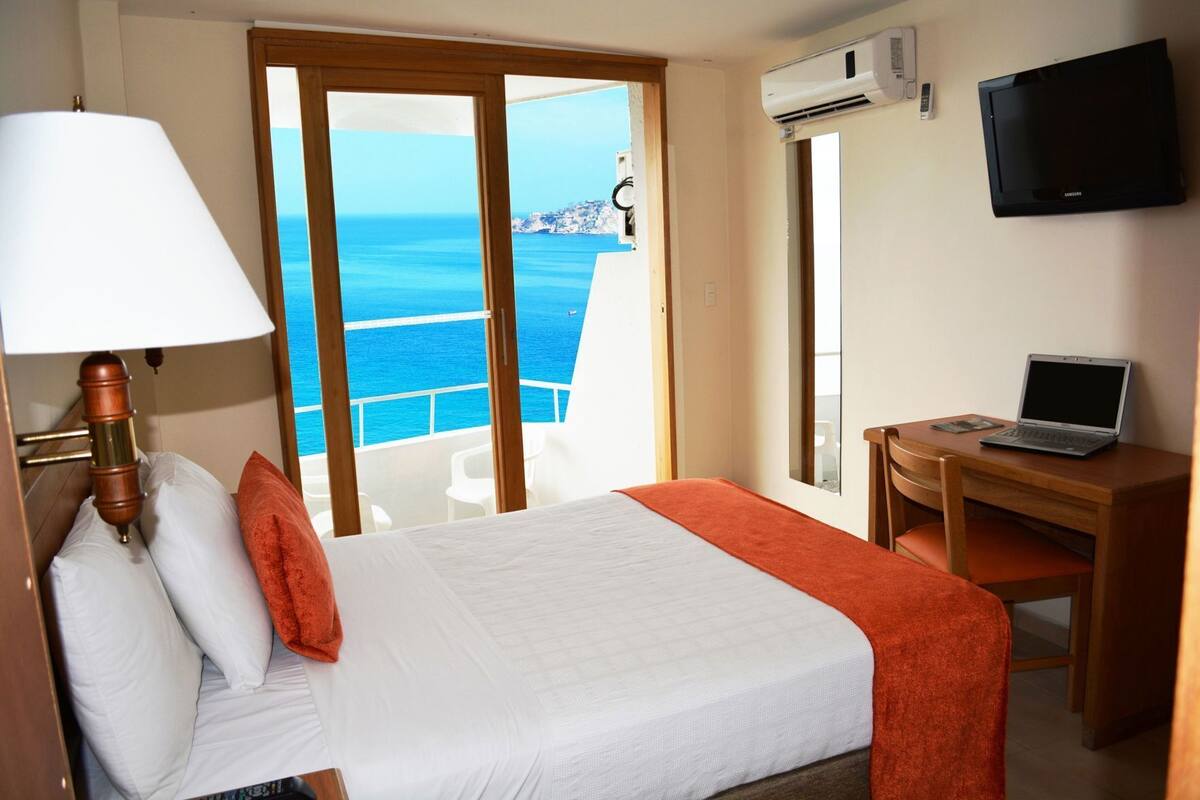 Une chambre d’hôtel avec un lit, un bureau avec un ordinateur portable, une télévision et une vue sur la mer.