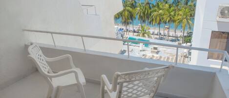 A balcony with two white chairs, a view of the beach with palm trees, and a pool.