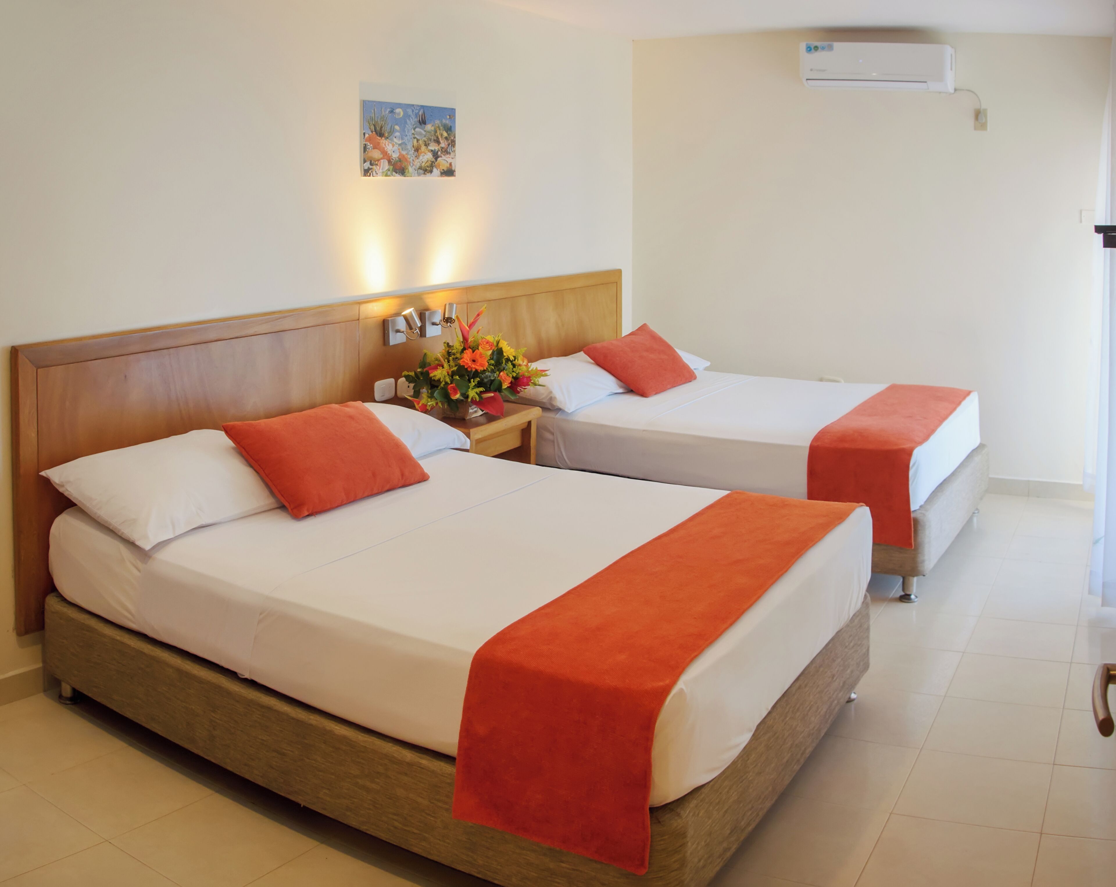 Two beds with wooden headboards, white bedding, orange pillows, a nightstand with a flower arrangement, and an air conditioning unit on the wall.