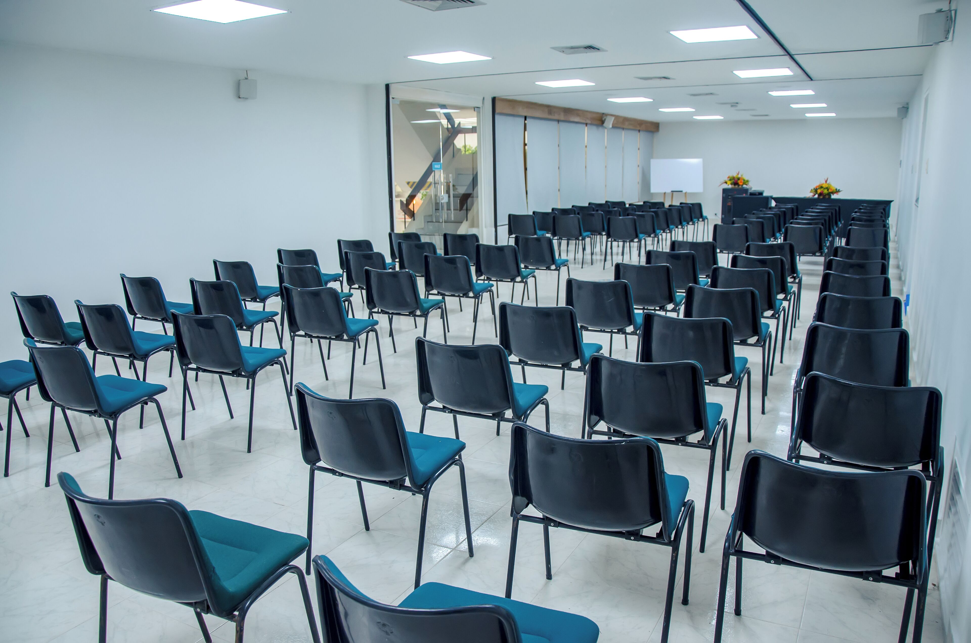 A conference room with rows of black chairs and teal cushions, a whiteboard, and a staircase in the background.