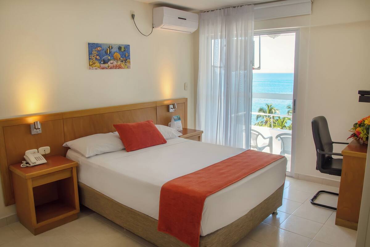 Une chambre d’hôtel avec un lit, un téléphone sur la table de chevet, une table de nuit, une chaise et une vue sur l’océan par la fenêtre.