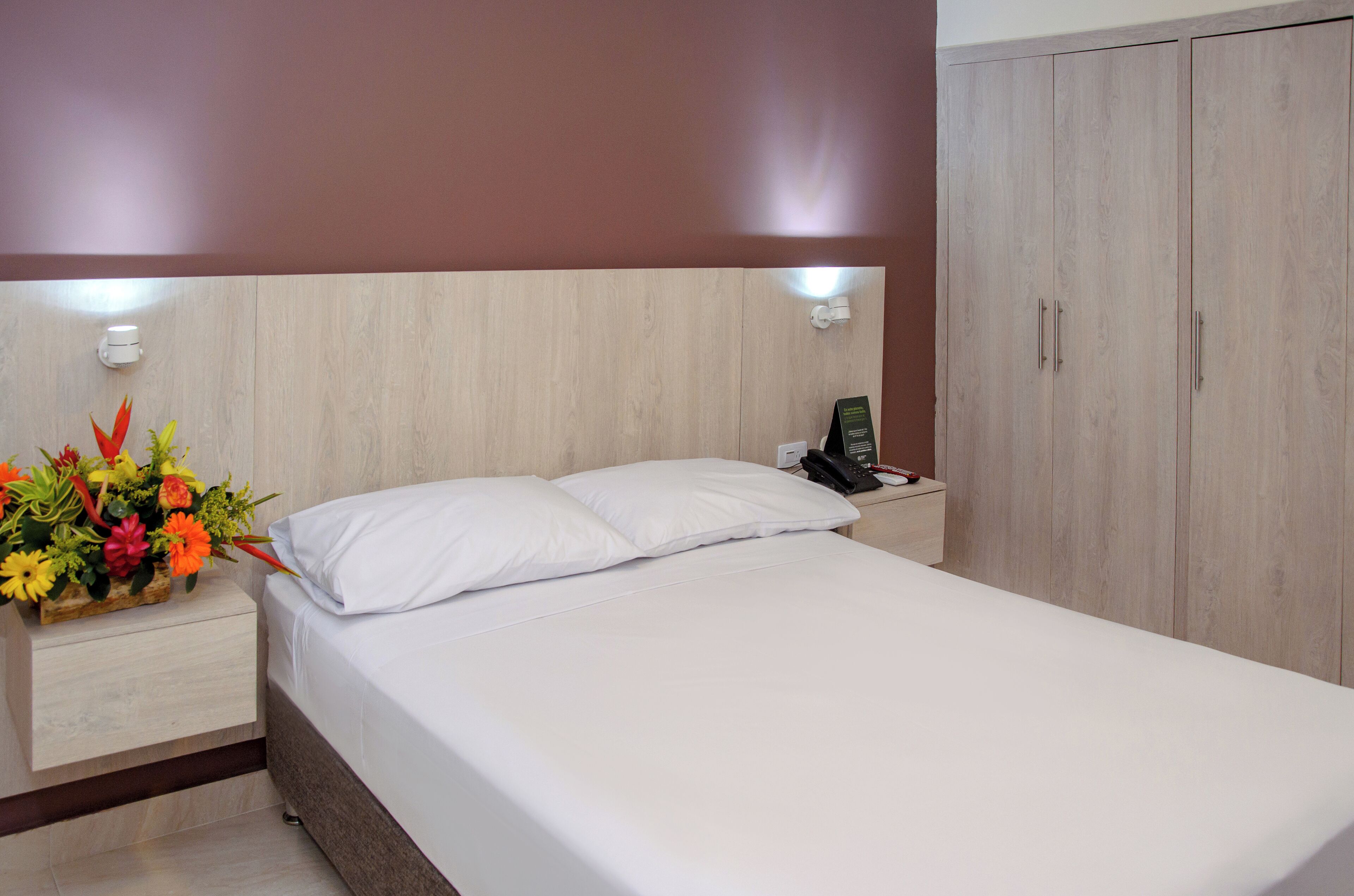 Een net opgemaakt bed met witte lakens, een nachtkastje met een telefoon en een bloemstuk, en een kledingkast met een licht houten afwerking.