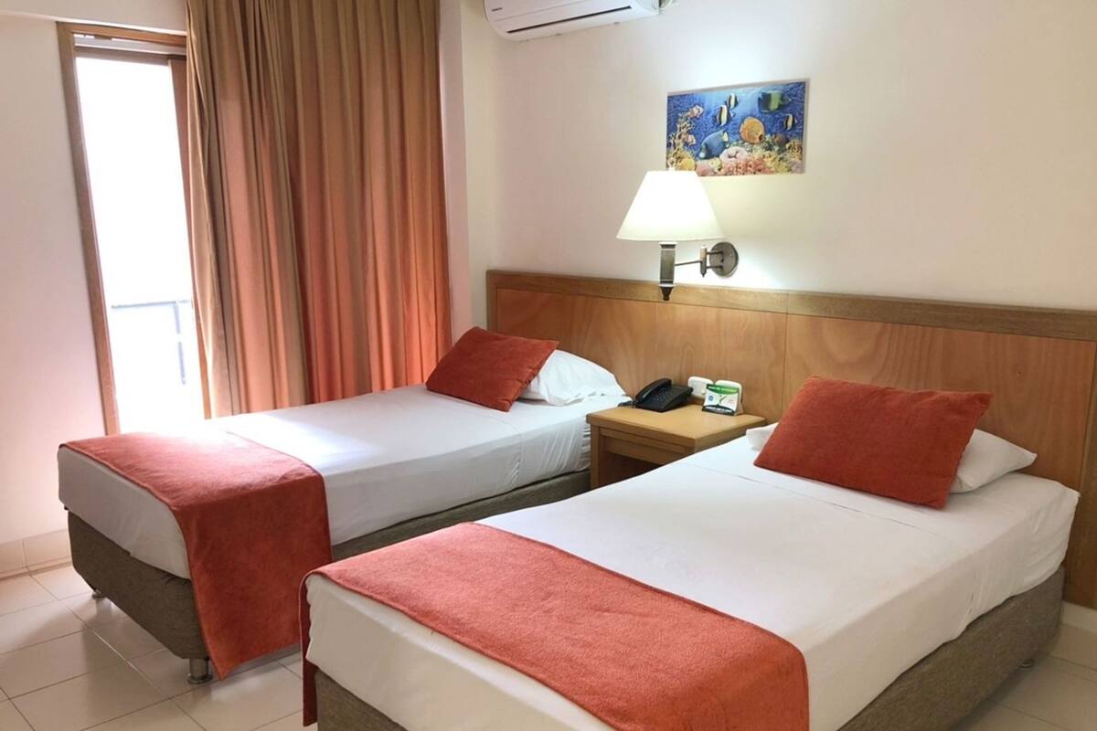 Une chambre d’hôtel avec deux lits, une table de chevet, une lampe, un tableau et une fenêtre avec des rideaux.