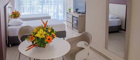 A hotel room with a bed, a TV, a dining table with chairs, and a flower arrangement.