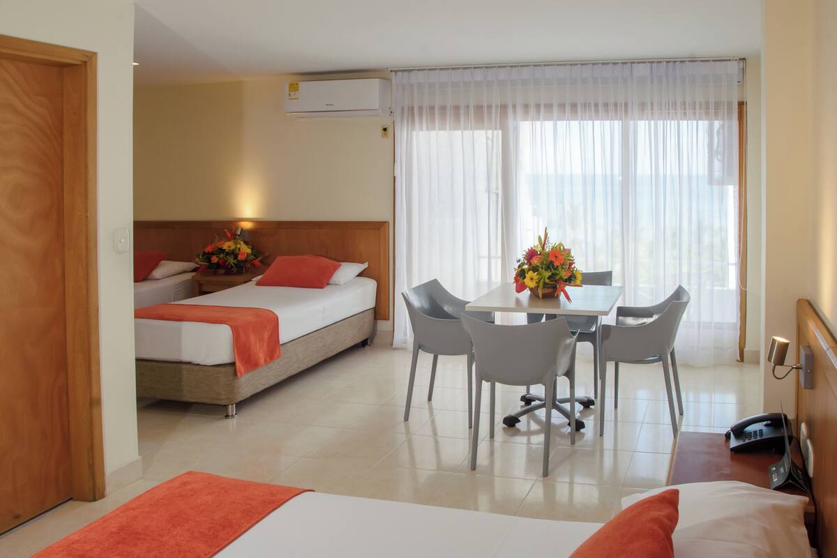 Une chambre d’hôtel avec deux lits, une table à manger avec des chaises, un téléphone et une fenêtre avec des rideaux légers.