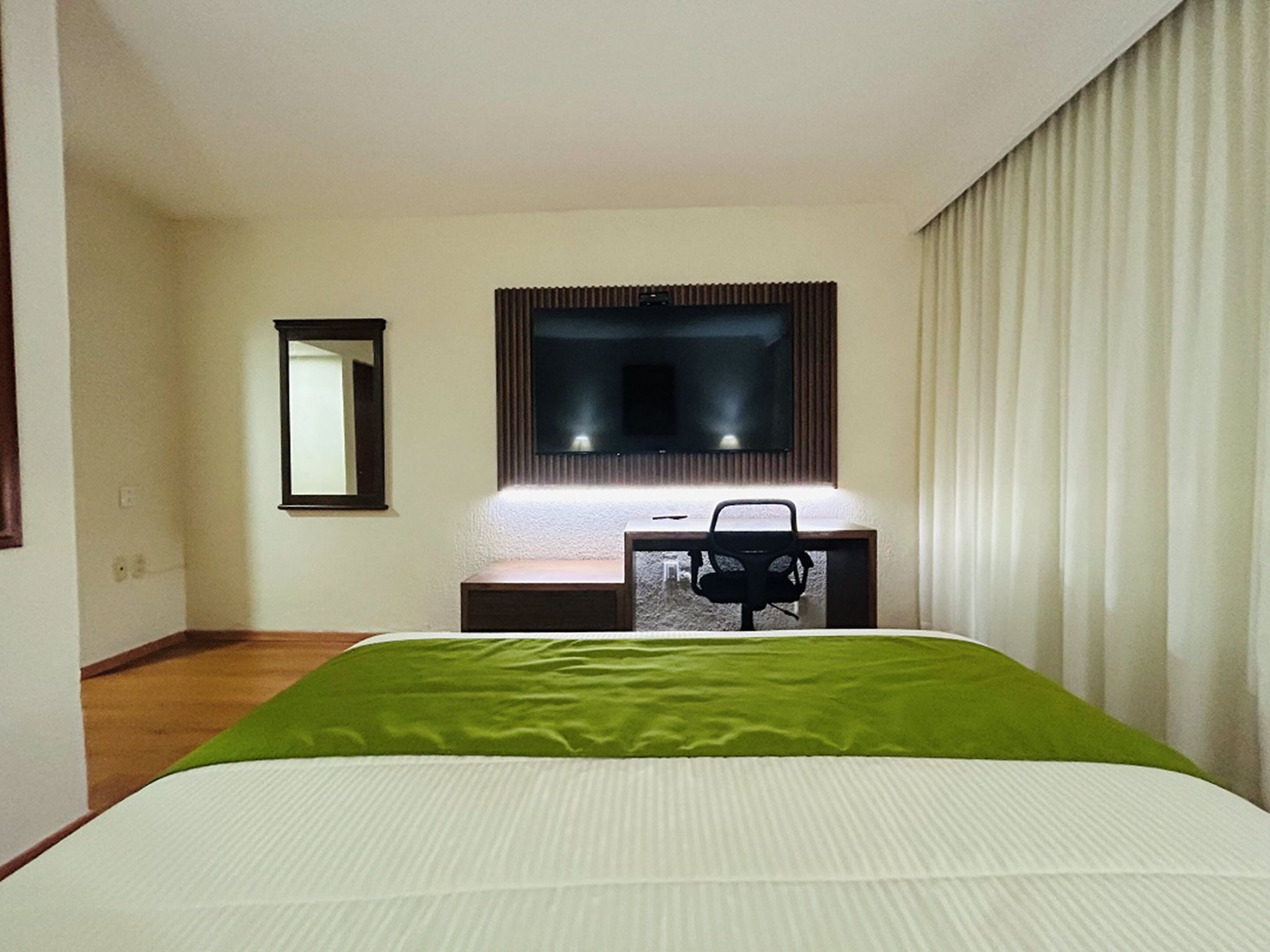 A hotel room with a bed, a desk with a chair, a television, and a window with curtains.
