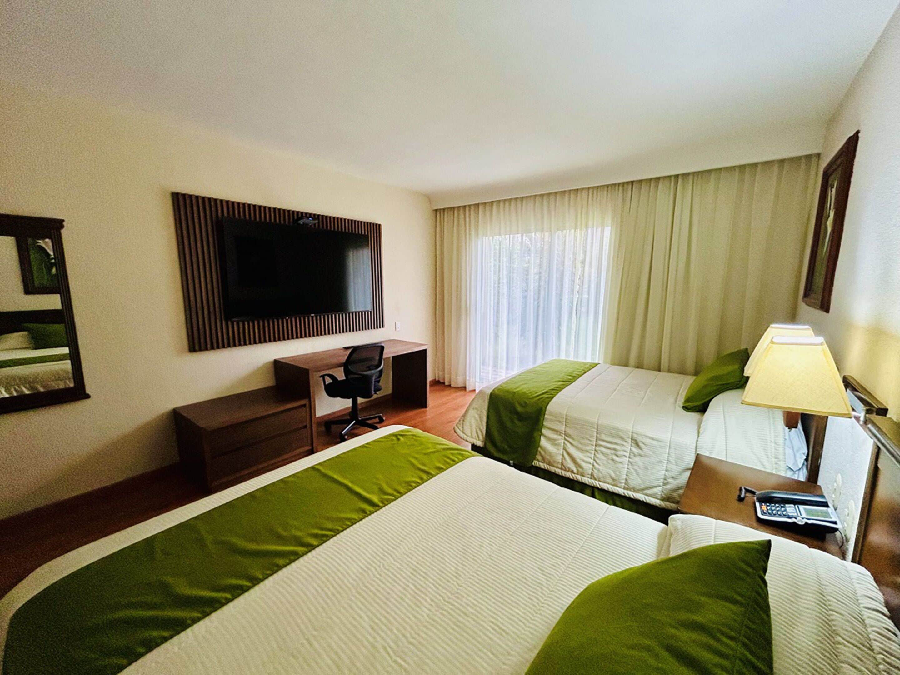 A hotel room with two beds, a desk, a chair, a television, and a lamp.