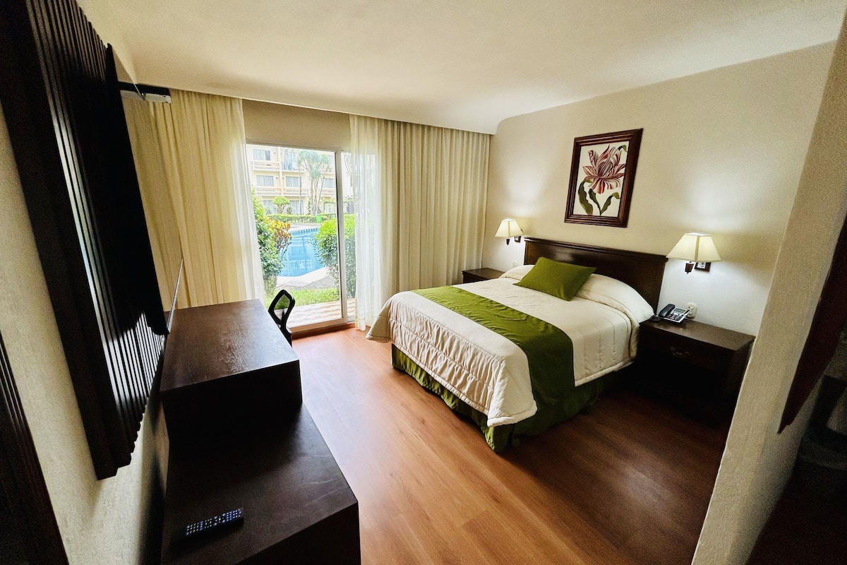 Ein Hotelzimmer mit Bett, Schreibtisch, Sessel, Fernseher, einem Fenster mit Vorhängen und einem Bild an der Wand.