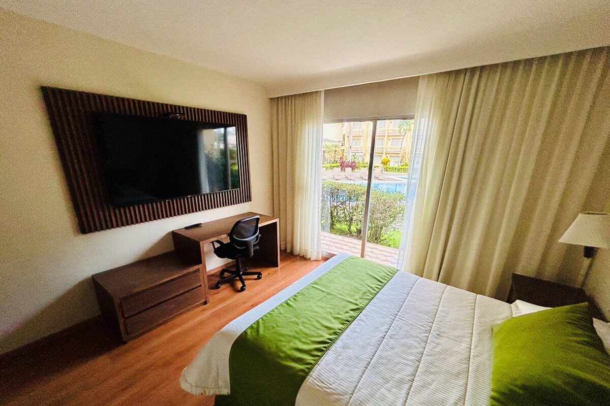 A hotel room with a bed, a desk, a chair, a TV, and a view of the outdoors.