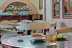 Interior de restaurante com mesas redondas, cadeiras de vime, área de bar ao fundo e um copo de vinho tinto sobre uma mesa.