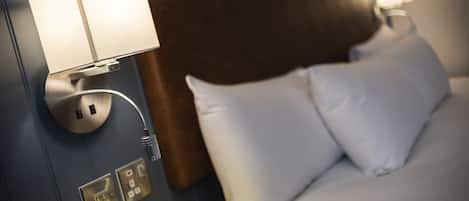 A hotel room with a bed, white pillows, a wooden headboard, and a wall-mounted lamp.