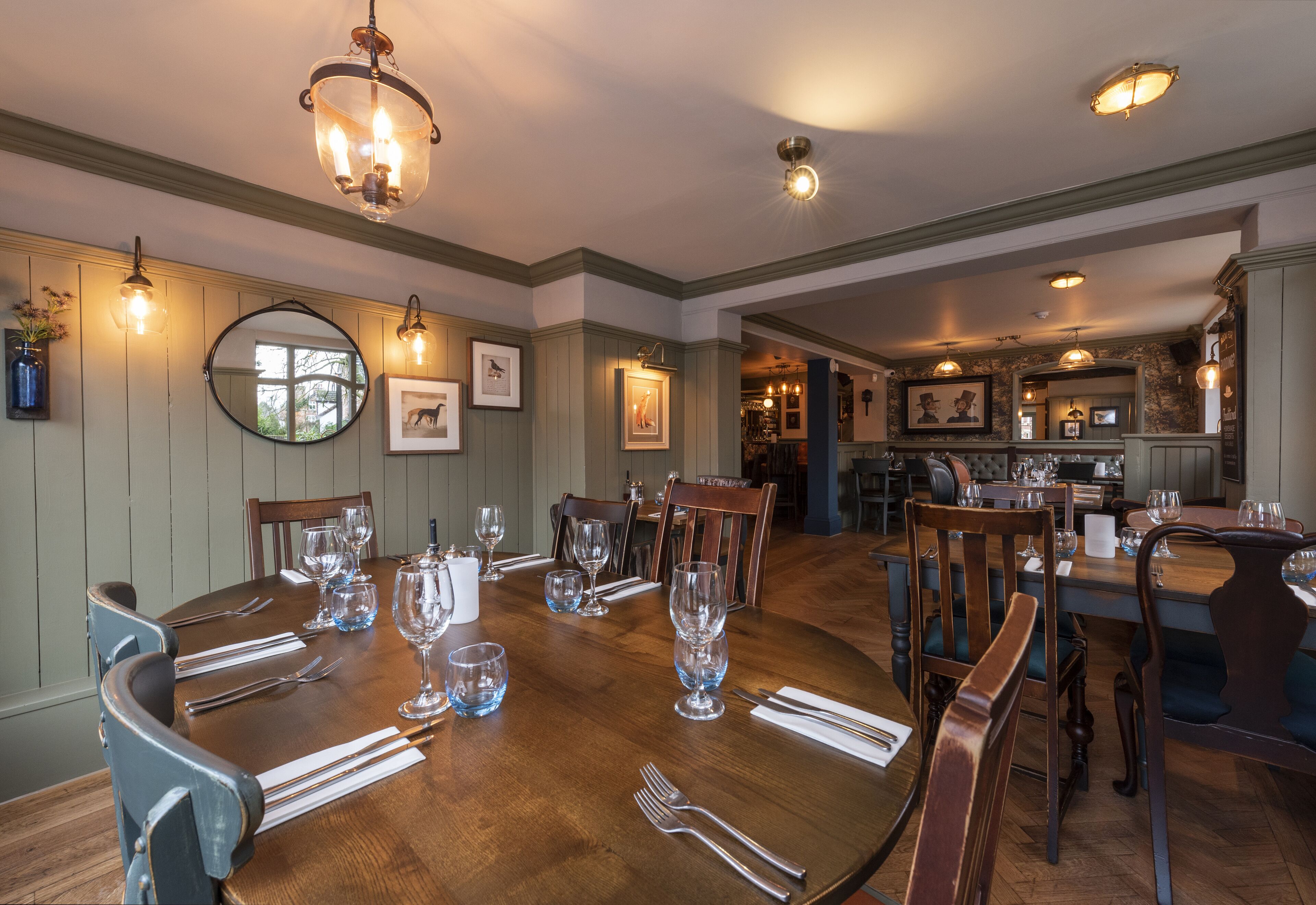 A dining room with a wooden table set for six, surrounded by chairs. There are framed pictures on the walls, a chandelier above the table, and a kitchen area in the background.