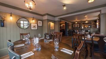 A dining room with a wooden table set for six, surrounded by chairs. There are framed pictures on the walls, a chandelier above the table, and a kitchen area in the background.