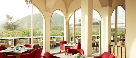 A dining area with red chairs and tables, large arched windows, and a view of greenery and hills.